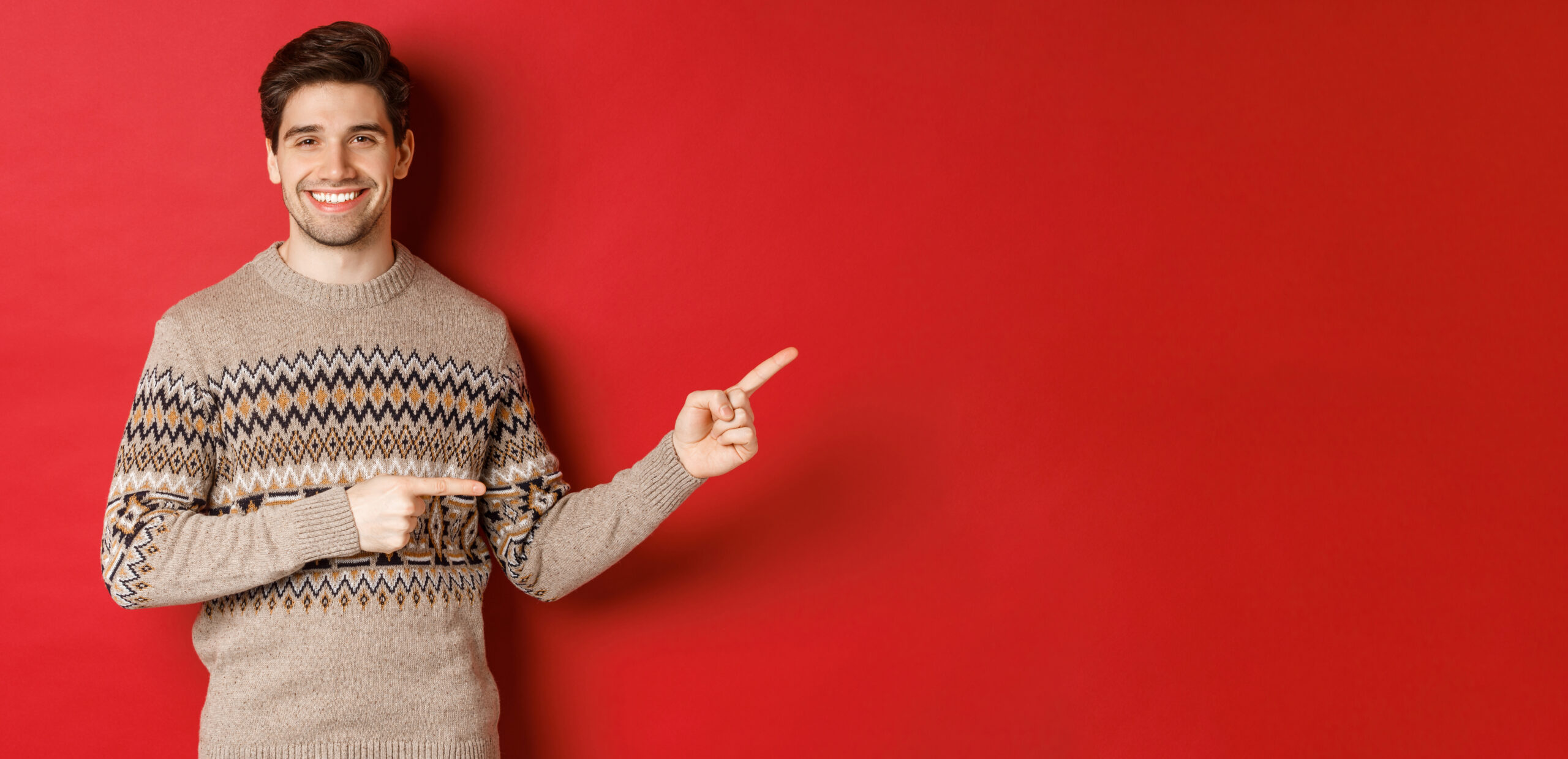 Image of attractive smiling man, wearing christmas sweater, pointing fingers right and showing new year advertisement, standing over red background.